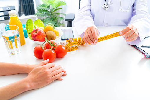Right nutrition and diet concept. Close up view of nutritionist hands holding a yellow tape measure explaining to patient the benefits of a healthy nutrition and lifestyle. An orange juice bottle, glass of water and fresh fruits and vegetables are on the doctor's desk and complete the composition. Copy space available for text and/or logo. High resolution 42Mp studio digital capture taken with Sony A7rII and Sony FE 90mm f2.8 macro G OSS lens