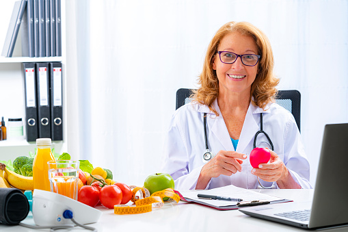 Right nutrition and diet concept. Smiling female doctor sitting at office desk holding a red heart shape explaining the benefits of a healthy nutrition and lifestyle. An orange juice bottle, glass of water, tape measure and fresh fruits and vegetables are on the doctor's desk and complete the composition. Copy space available for text and/or logo. High resolution 42Mp studio digital capture taken with Sony A7rII and Sony FE 90mm f2.8 macro G OSS lens