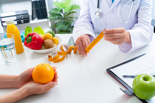 Right nutrition and diet concept. Close up view of nutritionist hands holding a yellow tape measure explaining to patient the benefits of a healthy nutrition and lifestyle. An orange juice bottle, glass of water and fresh fruits and vegetables are on the doctor's desk and complete the composition. Copy space available for text and/or logo. High resolution 42Mp studio digital capture taken with Sony A7rII and Sony FE 90mm f2.8 macro G OSS lens