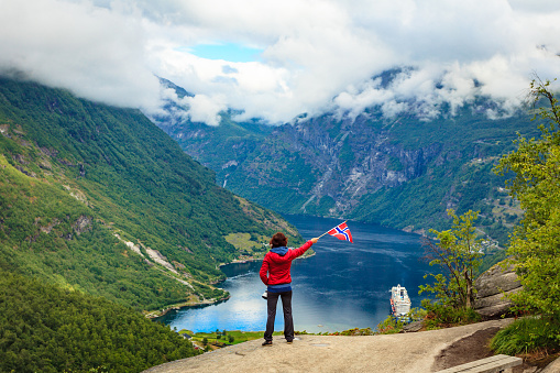 Female tourist enjoying scenic view over fjord Geirangerfjorden from Flydalsjuvet viewpoint, holding norwegian flag. Cruising vacation and travel.