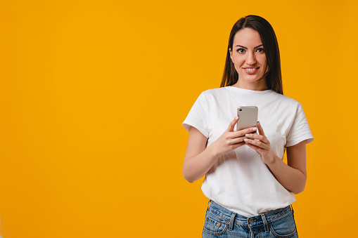 Smiling smart young caucasian woman using smart phone isolated over yellow background. Latin-american hispanic young female typing messages on cellphone, chatting, surfing the Internet