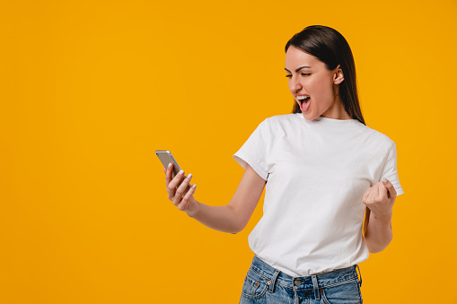 Happy as a winner excited young hispanic latin-american woman using smart phone isolated over yellow background. Emotional young caucasian female won the lottery big sum of money, huge success