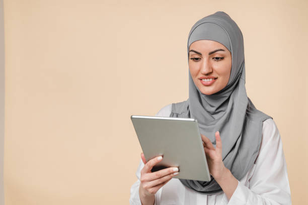 istock Smart intelligent islamic muslim arabian Middle Eastern young woman student using digital tablet isolated over beige background. Clever young female office worker in grey hijab chatting on tablet, sending messages, watching online webinars 1317480308