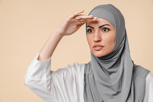 Young beautiful islamic middle eastern arabian muslim girl in grey scarf hijab looking far away in good future isolated over beige background. Hope, waiting for changes concept