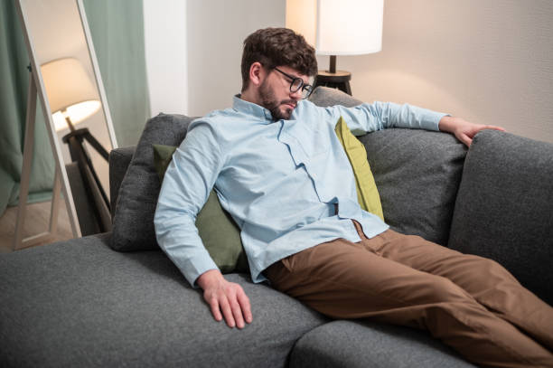 un jeune homme travaille de la maison, il est fatigué et s’allonge sur le canapé pour se reposer - lying down men isolated young adult photos et images de collection