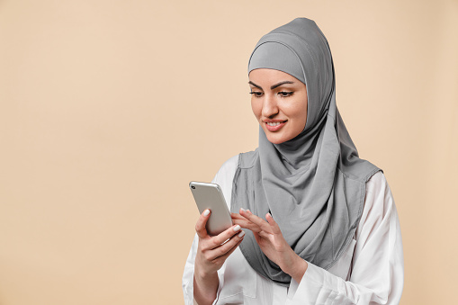Smiling smart young islamic middle eastern arabian muslim woman using smart phone isolated over beige background. Young female in grey hijab typing messages on cellphone, chatting, surfing the Internet