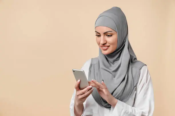 Photo of Smiling smart young islamic middle eastern arabian muslim woman using smart phone isolated over beige background. Young female in grey hijab typing messages on cellphone, chatting, surfing the Internet