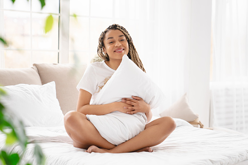 Young african-american woman hugging her comfy pillow sitting on the bed with white linens and orthopedic mattress, relaxing before starting a new day