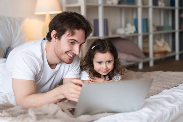 istock Caucasian father and daughter using laptop looking at screen choosing goods online lying on the bed, cheerful dad talking to child showing new computer watching video, doing internet shopping together 1317492888
