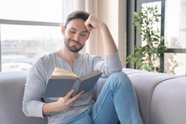 istock Excited relaxed young caucasian man reading a book, e-learning, having a break after work at home. Leisure time, weekend and hobby indoors. Adult student 1317495689