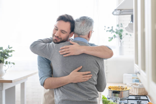 fils caucasien adulte affectueux affectueux affectueux embrassant étreindre son vieux père aîné âgé dans la cuisine tout en cuisinant le déjeuner, le dîner, préparant le repas ensemble. bonne fête des pères ! je t’aime, papa ! - parent denfant adulte photos et images de collection