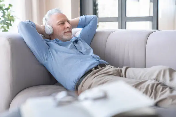 Relaxed old elderly senior mature man grandfather listening to the music in headphones earphones, having rest on the sofa at home. Active seniors concept