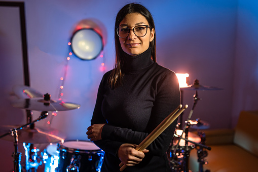 Front view portrait of adult caucasian woman posing in front of drums set in dark room or studio - Drummer woman standing looking to the side at night - copy space real people