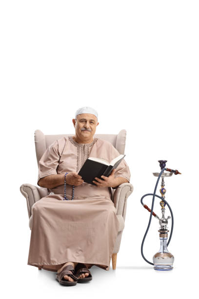 Muslim man sitting in an armchair with reading a book and a shisha next to him Muslim man sitting in an armchair with reading a book and a shisha next to him isolated on white background middle eastern ethnicity mature adult book reading stock pictures, royalty-free photos & images