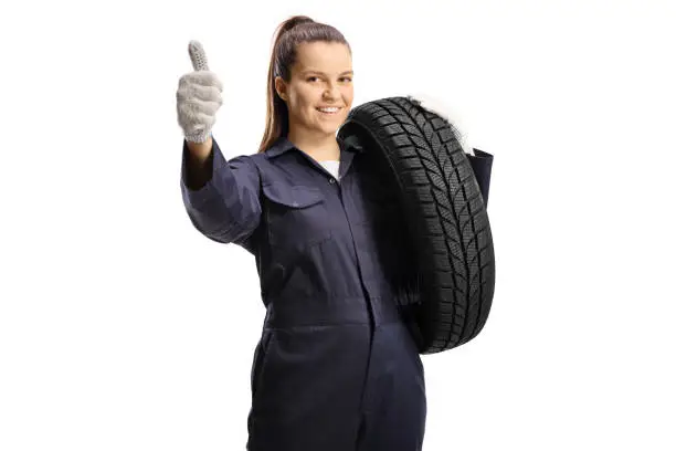 Woman mechanic worker carrying a car tire and gesturing a thumb up sign isolated on white background