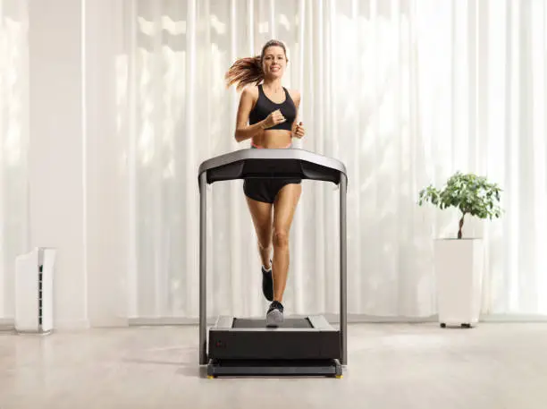 Photo of Young woman in shorts and top running on a treadmill at home