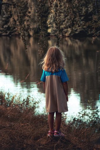 Lonely child girl standing alone in forest on river bank and looking into the distance in countryside concept for sadness and loneliness