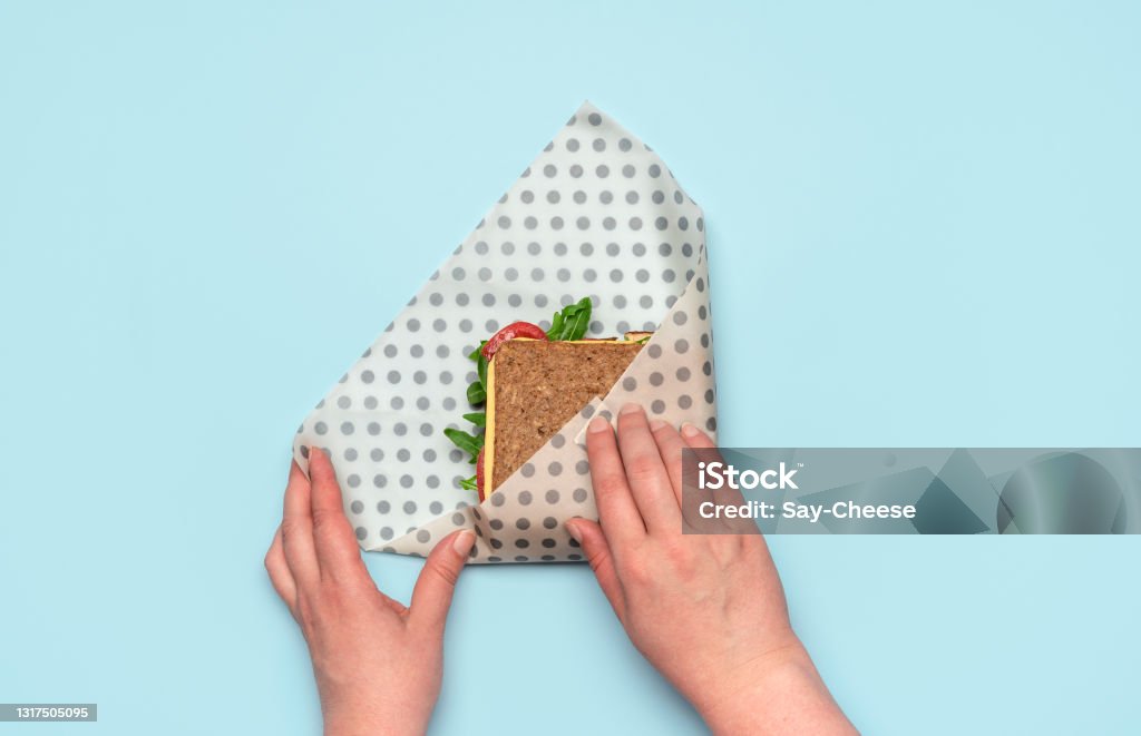 Wrapping sandwich in a beeswax cloth, top view. Ethical consumerism. Woman's hands wrapping a sandwich in a beeswax cloth on a blue table, above view. Healthy sandwich with wholemeal bread and vegan ingredients, wrapped in reusable beeswax paper. Wrapping Paper Stock Photo