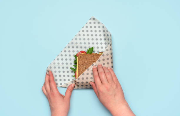 Wrapping sandwich in a beeswax cloth, top view. Ethical consumerism. Woman's hands wrapping a sandwich in a beeswax cloth on a blue table, above view. Healthy sandwich with wholemeal bread and vegan ingredients, wrapped in reusable beeswax paper. beeswax photos stock pictures, royalty-free photos & images