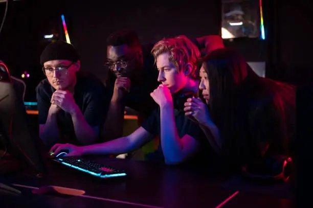 Photo of Two guys and a girl in medical masks in a computer club give support to their friend during his game