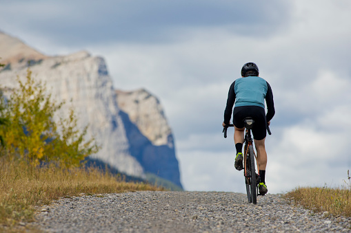 A man goes for a gravel road bicycle ride at the Canmore Nordic Centre in Alberta, Canada. Gravel bicycles are similar to cyclocross bikes with strong wheels and tires for riding on rough terrain. He is wearing a bicycle helmet, sunglasses, and road cycling clothing.