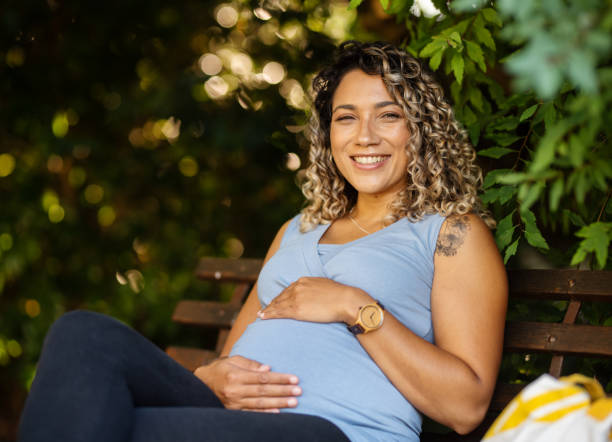 Smiling young pregnant woman sitting outdoors on a park bench Portrait of a smiling young pregnant woman cradling her tummy while sitting outside on a park bench in the summer pregnant stock pictures, royalty-free photos & images