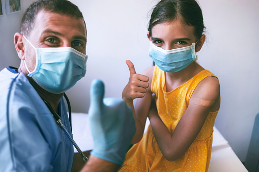 Happy little girl with adhesive bandage on her hand taking a picture with a doctor after COVID-19 vaccine.