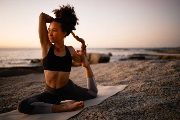 full length shot of an attractive young woman practicing yoga on the beach at sunset - stretch beach imagens e fotografias de stock