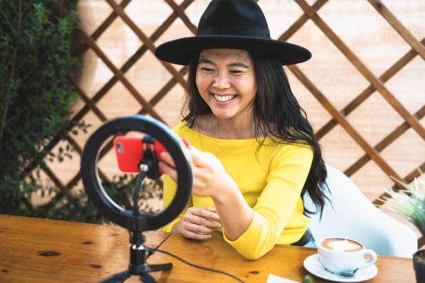 Happy Asian woman vlogging online using smartphone cam and influencer led while drinking a coffee sitting in bar