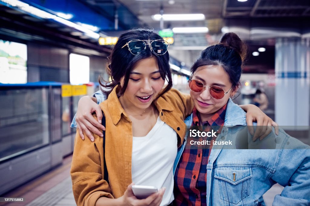 Females discussing over phone at subway station Young women looking in mobile phone. Smiling female friends are exploring city in weekend. They are at subway station. Subway Stock Photo