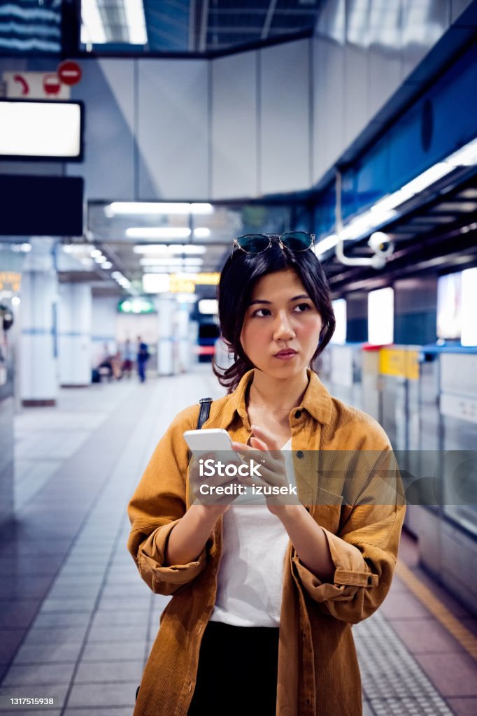 Female with mobile phone waiting at subway station Young woman with cellphone looking away. Female in casuals is waiting for train. She is at subway platform. Females Stock Photo