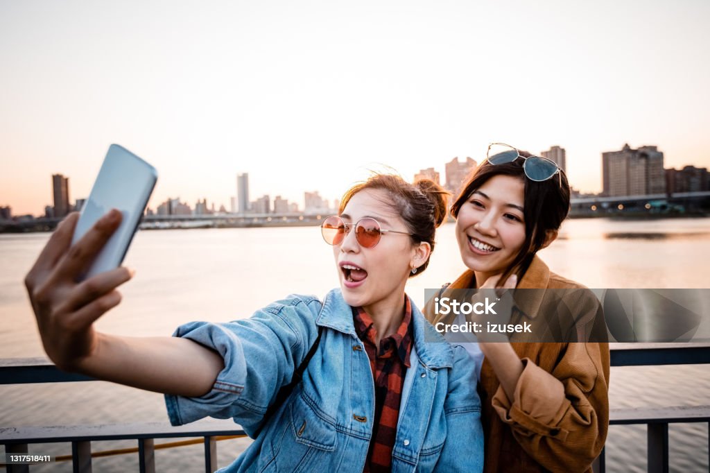 Happy women taking selfie during city break Carefree young women taking selfie through smart phone. Female friends are enjoying weekend at sunset. They are on city break. Selfie Stock Photo