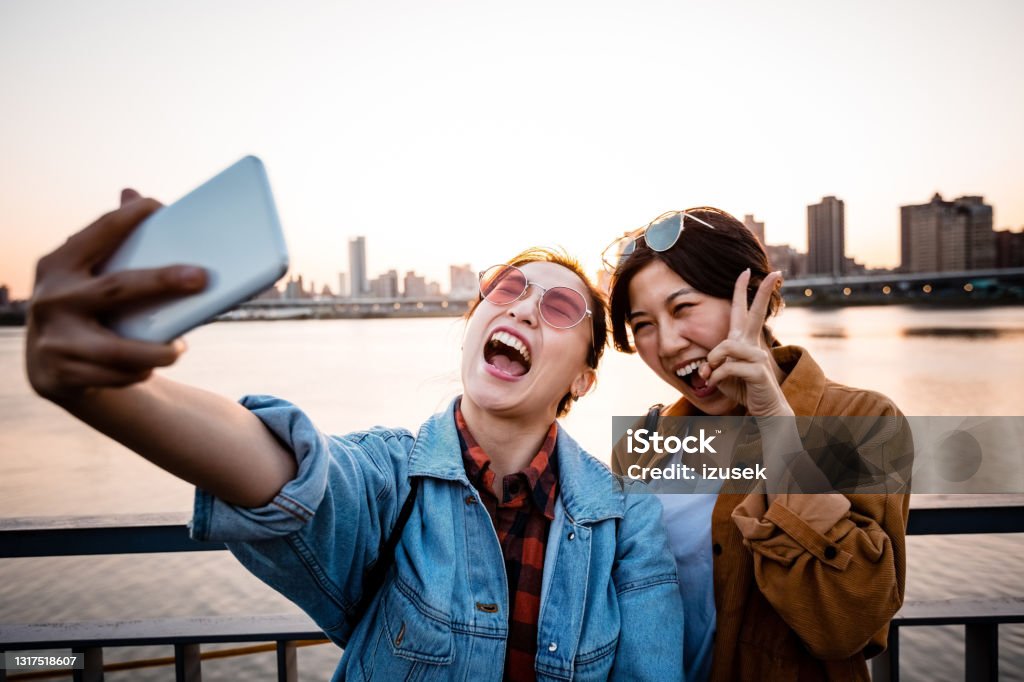 Cheerful women taking selfie during sunset Happy female friends taking selfie through smart phone. Young women are enjoying weekend together at sunset. They are wearing casuals. 20-24 Years Stock Photo