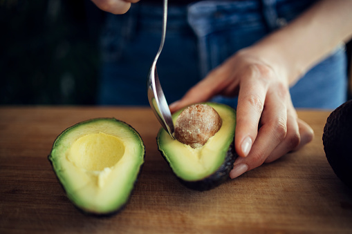 Unrecognizable female person peeling perfectly ripe avocado with spoon on wooden cutting board in kitchen