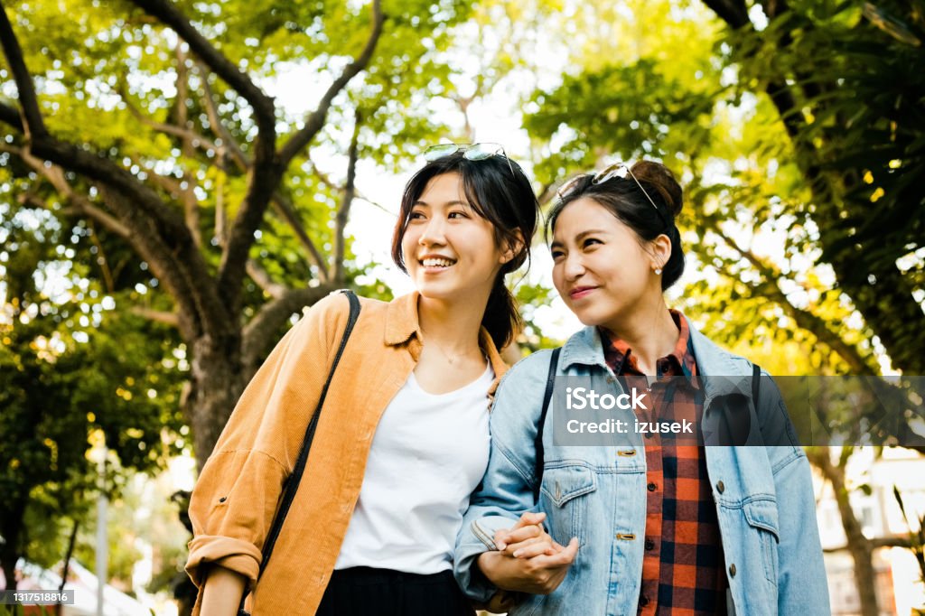 Young female friends in park during weekend Smiling women looking away while standing against trees. Happy young female friends are holding hands. They are in public park. 20-24 Years Stock Photo