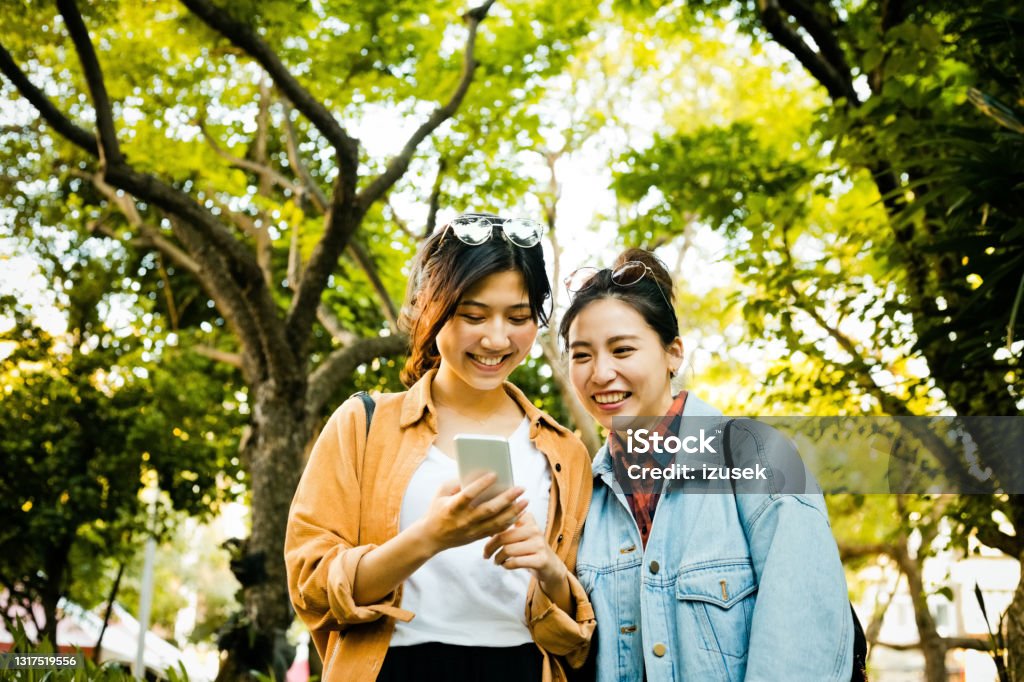 Happy female friends in park during weekend Smiling young women looking in smart phone. Happy female friends are enjoying weekend together. They are in park. 20-24 Years Stock Photo