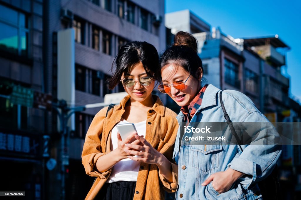 Female friends exploring city during sunny day Women looking in mobile phone while exploring city. Young female friends are spending weekend together. They are wearing casuals. Smart Phone Stock Photo