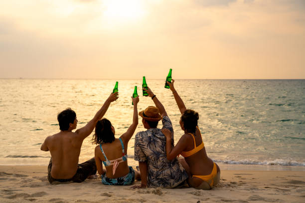 ハッピーアジアの男女の友人のグループは、夏の日没時にビーチで一緒に話してビールを座って飲んでいます。 - friends on the beach ストックフォトと画像