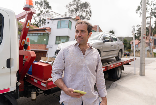 Latin American man having a vehicle breakdown and looking worried while a tow truck takes his car to the auto repair shop - roadside assistance concepts