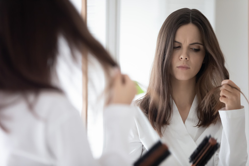Worried girl concerned about hair loss, combing at mirror, holding hair brush and lock. Upset young woman counting fell out hairs. Haircare, health problem concept. Mirror reflection head shot