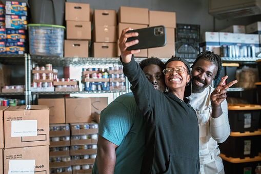 A group of young adult friends volunteer time working at a food bank, processing donations of packaged food products and clothing.  They pause from their work to take a selfie with their smartphone.