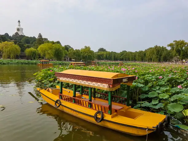 A yellow boat parked at the shore of a small lake in Beihai Park, Beijing, China. There is a white Beihai Pagoda on the hill. Lots of water lilies. Thick smog above the city. Air pollution.