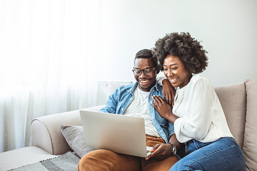 Young black couple sit on the sofa using laptop. Black couple using laptop at home look at each other. Overjoyed african American couple sit relax on cozy couch look at laptop screen triumph winning lottery online