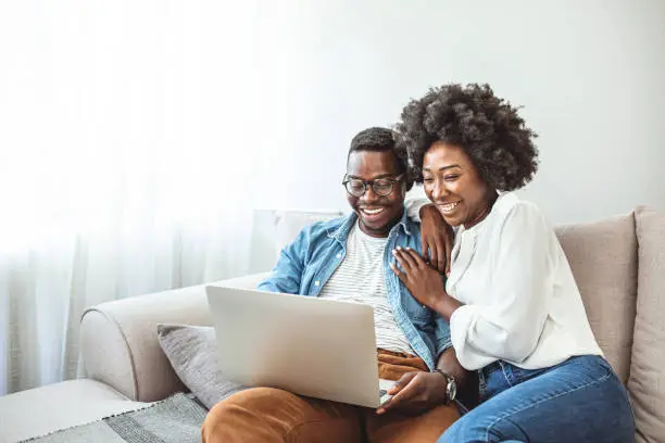Photo of Black couple using laptop at home look at each other.