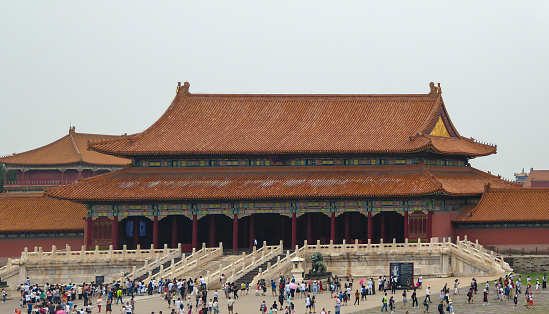 A view on a big square inside a building complex of Forbidden City in Beijing, China. The buildings have very richly decorated rooftops, with elements of gold. Thera are lots of people on the square.