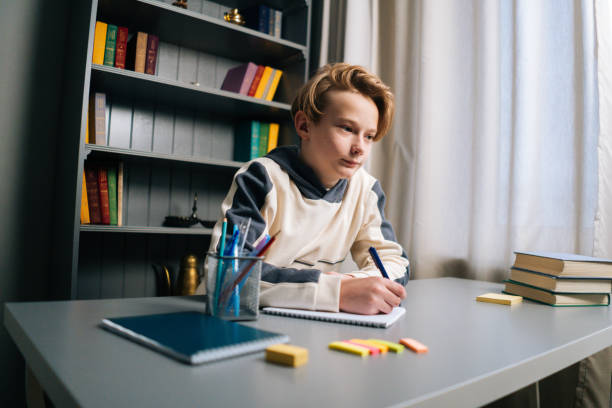 istock Low-angle view of thinking pupil boy thoughtful writing in notebook with pen sitting at desk near window. 1317565393