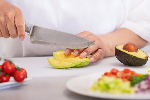 Hands using a kitchen knife to slice an avocado half on a cutting board. Selective focus and close up. Cooking and healthy food concept.