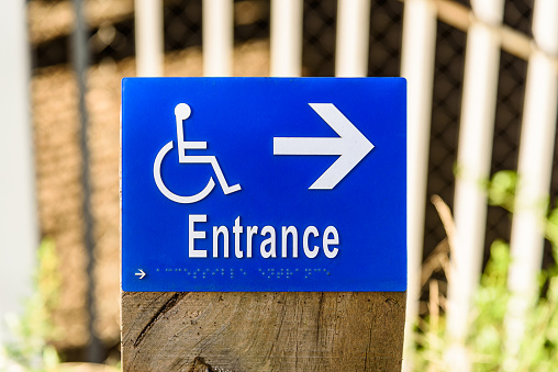 A blue sign on a wooden post featuring wheelchair and arrow indicating the direction to an accessible entrance