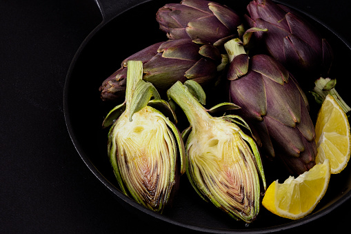 Uncooked organic purple artichokes with lemon slices on a frying pan. Rustic style on black surface. Dark photography style with copy space