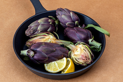Uncooked organic purple artichokes with lemon slices on a black cooking pan. Rustic style on brown surface. Top view with copy space